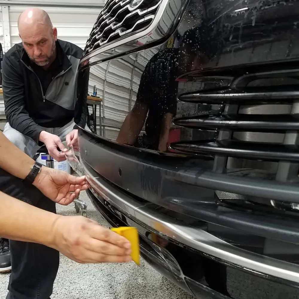 Two technicians using a yellow squeegee to apply paint protection film on a bumper.
